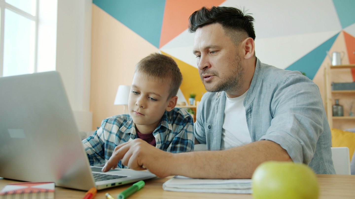 Man teaching boy to use laptop at table with school supplies.
